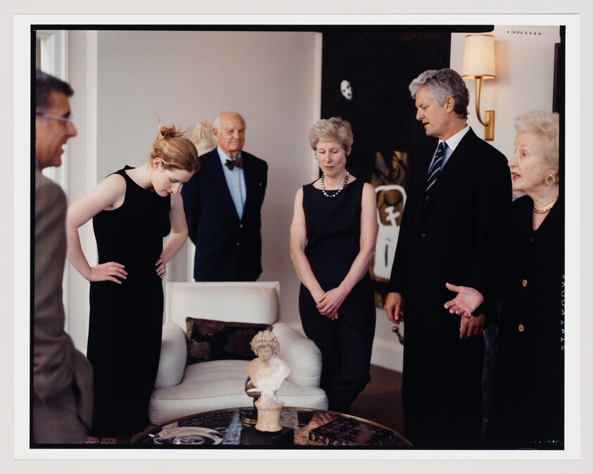 Several people in formal clothing stand around and examine a small bust sculpture on a table.