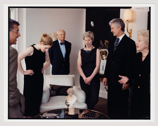 Several people in formal clothing stand around and examine a small bust sculpture on a table.