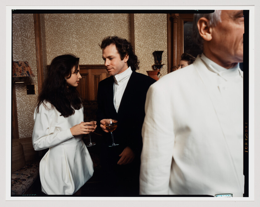 A young couple in formal wear talk while holding wine glasses at a social gathering.