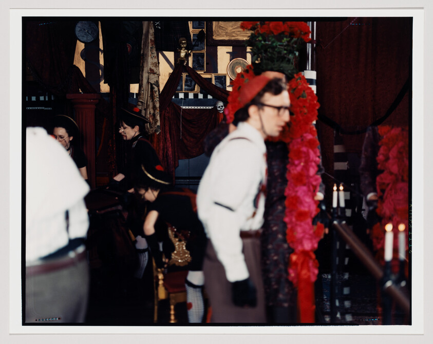 A man in vintage costume walks past a tall red floral wreath while others sit nearby.