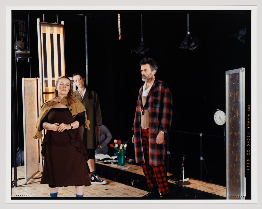 Three actors on a stage with a theatrical set, including a woman in the foreground wearing a brown dress and a shawl, a woman in the background in a green coat, and a man on the right in a plaid robe. The stage is adorned with props like a clock, hanging shoes, and a tiled floor, creating an intimate performance atmosphere.