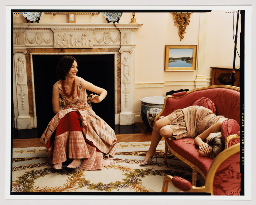 Two women in vintage dresses laugh and rest in an ornate living room.