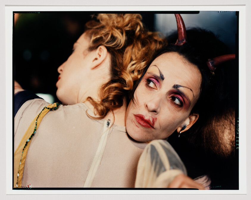 Woman wearing devil horns and theatrical makeup embraces another person while looking away.
