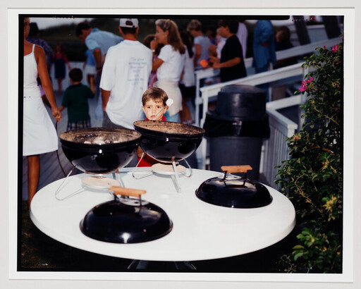 A young boy stands behind tabletop grills at a crowded outdoor gathering, staring toward the camera.