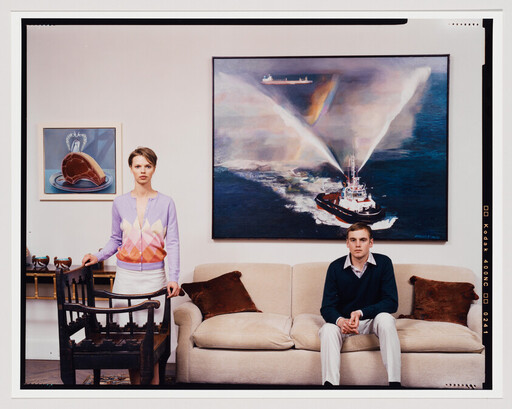 Young woman stands by a chair while a young man sits on a sofa beneath a large ship painting.