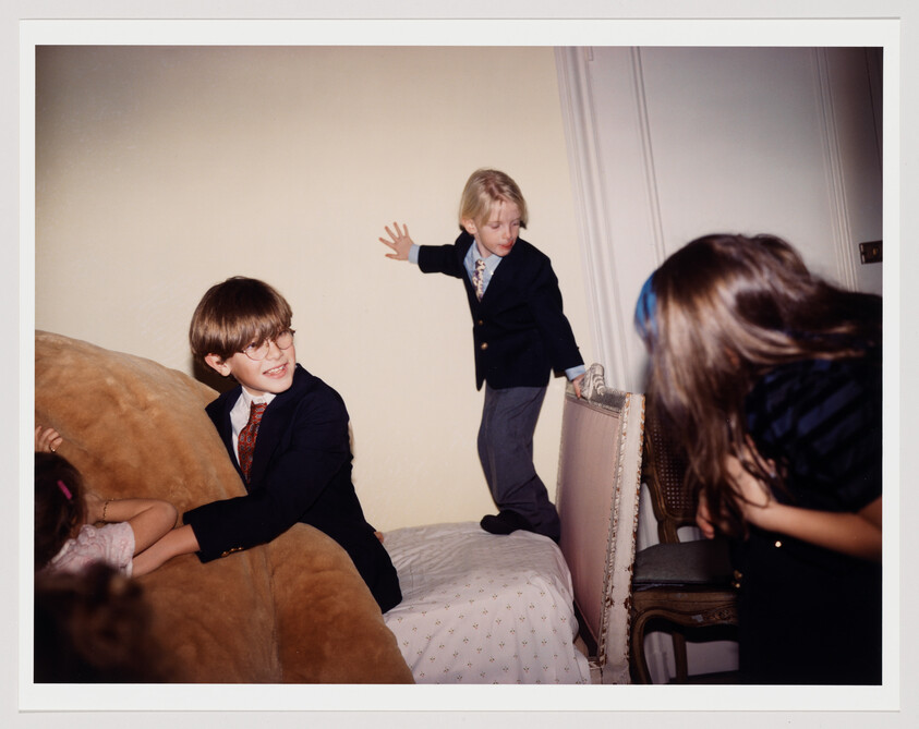 Three children in suits and dresses play energetically on a bed, one boy balancing on the headboard.