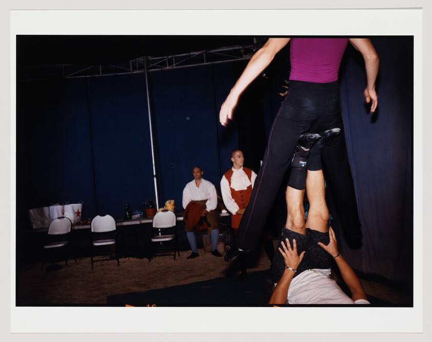 A performer is being held upside down by another person's hands, with only their legs and torso visible in the frame. In the background, two individuals are seated, one in a red and white costume, and the other in casual attire, both observing the scene. The setting appears to be a backstage area with blue curtains and chairs along a table with various items on it.