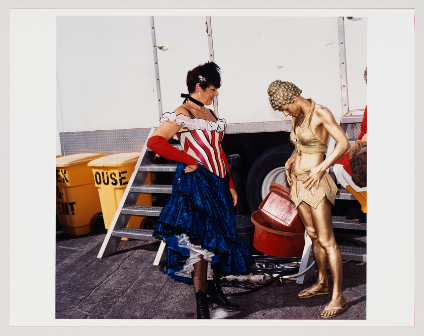 Two performers in elaborate costumes stand near a white trailer and yellow bins labeled "EXCLUSIVE AGENT." The person on the left is dressed in a patriotic-themed outfit with a blue skirt and red and white stripes, while the person on the right wears a golden costume resembling ancient Roman armor. They appear to be engaged in a conversation behind the scenes of an event or performance.