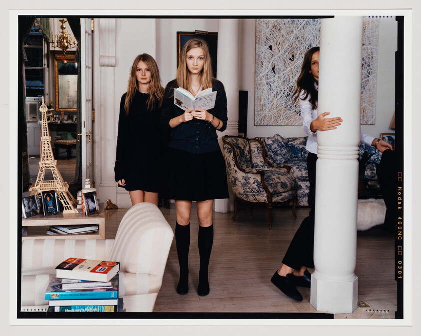 Two young women stand in an elegantly decorated room with a large white column between them. The woman on the left is dressed in a black long-sleeve dress and black knee-high socks, with her hair down, looking directly at the camera. The woman on the right, holding an open book, wears a dark blazer over a white blouse paired with a black skirt and similar knee-high socks, her gaze directed towards the book. A model of the Eiffel Tower, books, and a striped sofa are in the foreground, while ornate furniture and an intricate wall art piece adorn the background.