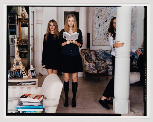 Three teenage girls in a living room, one reading a book while another hugs a pillar.