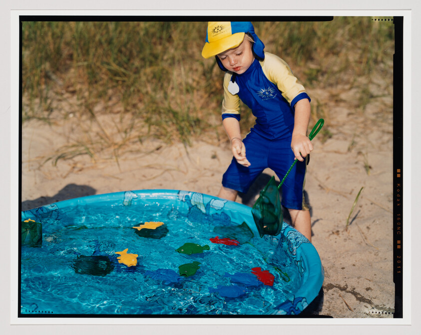 Young child in blue swimsuit uses a net to scoop toy fish from a small pool.