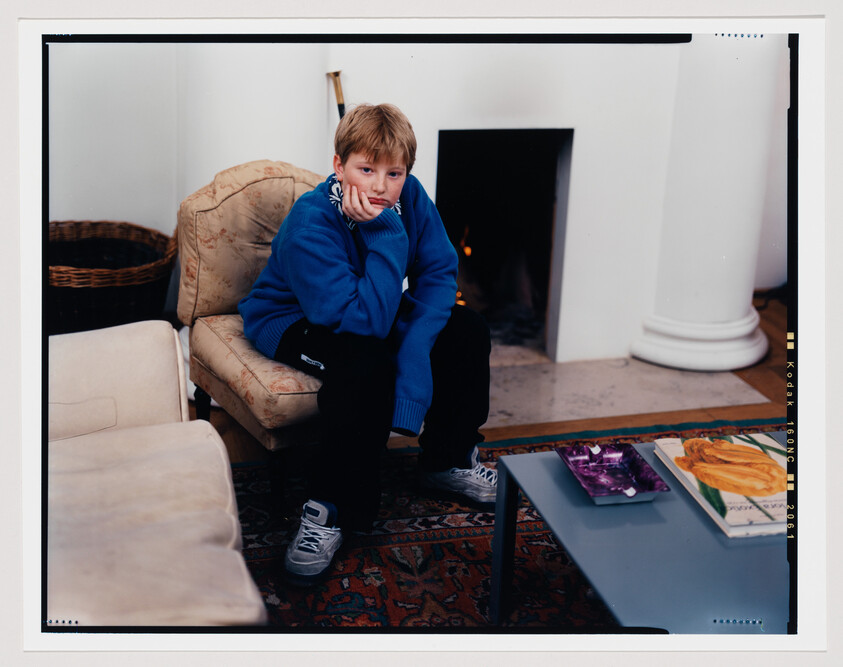 A young boy with light hair sits on his heels on a blue coffee table, resting his chin on his hand, looking pensively at the camera. He wears a bright blue sweater and dark pants. Behind him is a cream-colored armchair and a fireplace with a lit fire. On the table in front of him are two magazines with colorful covers. The room has a warm ambiance with a patterned rug on the floor. The image has a white border with black and yellow markings resembling those of a film negative, including the text "Kodak -- 160NC".