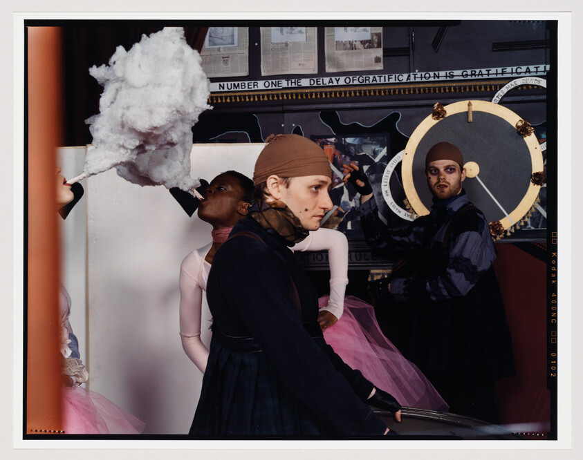 A staged photograph featuring three individuals in a storefront window display. The person on the left appears to be blowing a large cloud of cotton resembling smoke, while the person in the center, dressed in a headscarf and skirt, gazes intently to the side. The person on the right, wearing a cap and a patterned jacket, holds a circular object with a pendulum, looking directly at the camera. The backdrop includes text and graphics, creating an artistic and surreal scene.