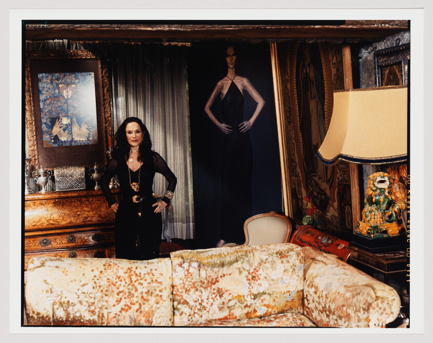 A woman stands confidently in an opulent room decorated with rich textiles and antique furniture. She is dressed in a black outfit with sheer sleeves and a prominent gold belt. Behind her, a large framed painting and a life-sized portrait of a model in a black dress add to the luxurious ambiance. The room is adorned with heavy drapes, a floral-patterned sofa, and a classic lampshade, creating a sense of vintage elegance.