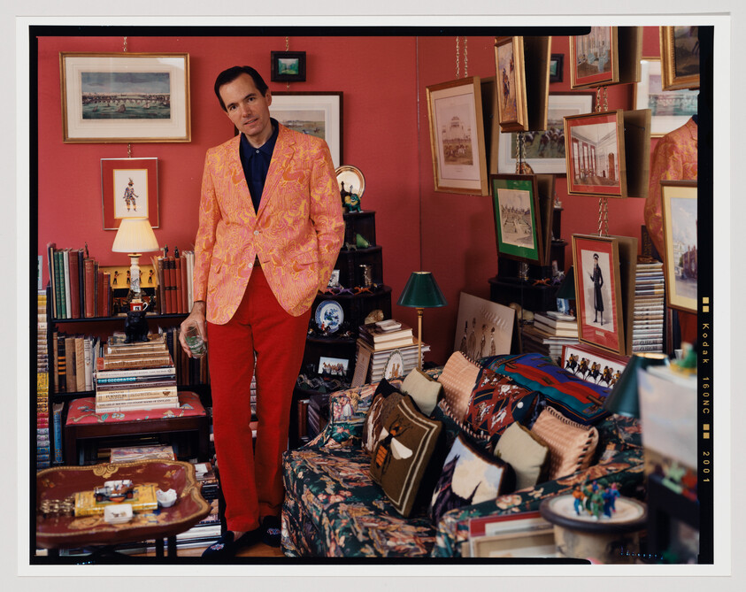 A man in a pink patterned jacket and red pants stands holding a glass in a cluttered living room.