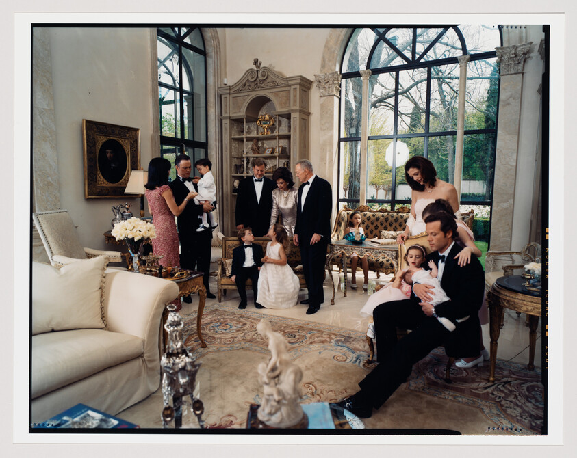 A staged family portrait in an opulent room with large windows, featuring multiple generations dressed in formal attire. Some adults are standing and conversing, while others are seated with children on their laps. The room is decorated with antique furniture, ornate decorations, and framed artwork.