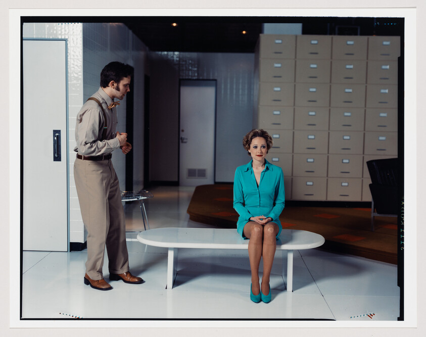 Woman in teal dress sits on a bench while a man in beige talks to her.