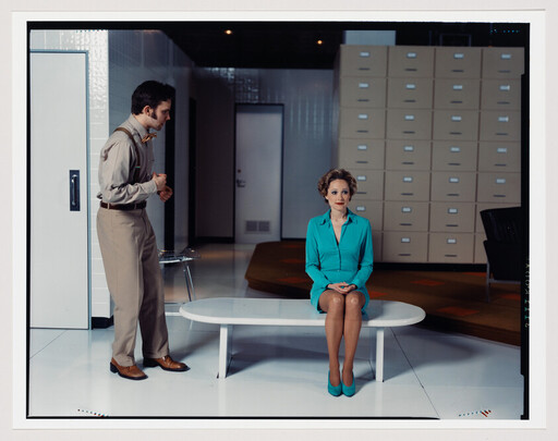 Woman in teal dress sits on a bench while a man in beige talks to her.