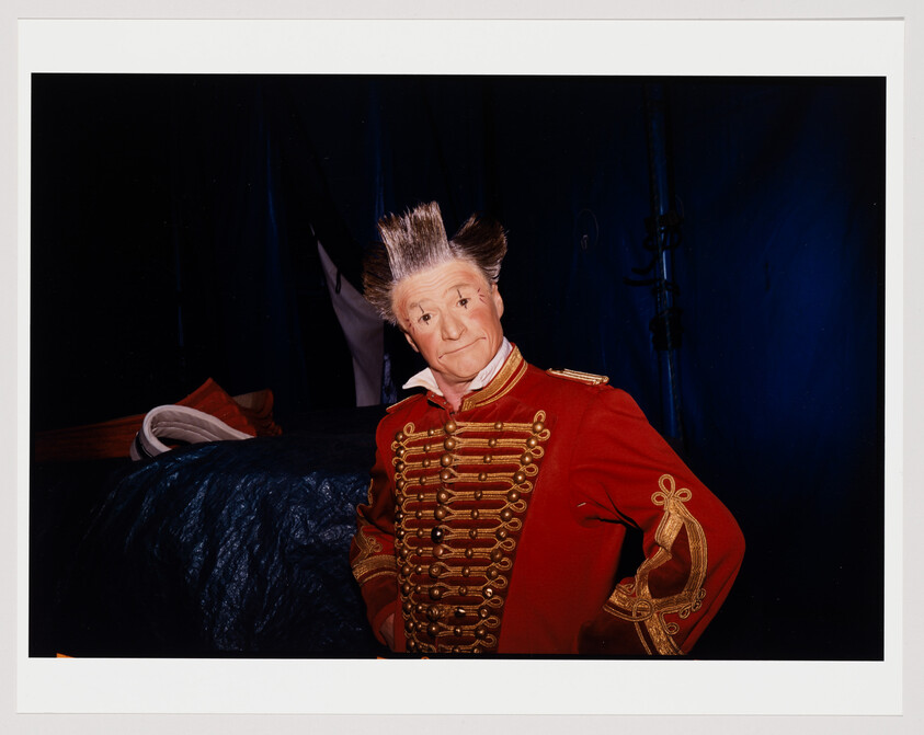 A person wearing a red military-style jacket with gold embellishments stands in front of a dark blue backdrop. The individual has a distinctive hairstyle with the hair standing upright and a somewhat exaggerated facial expression with raised eyebrows.