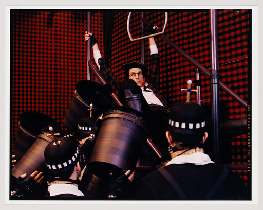 A performer hangs from scaffolding while costumed crew lift large black drums below him.