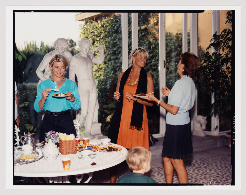 Three women stand by a table of snacks, smiling and talking while holding plates.