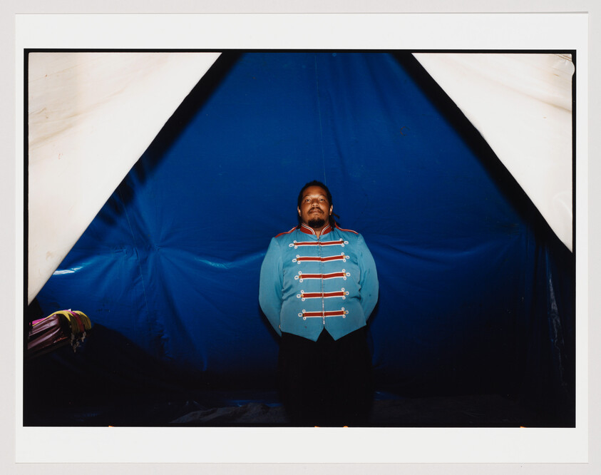 A person in a blue and red uniform with decorative buttons stands in front of a blue and white tent backdrop, giving the impression of a circus or performance setting.