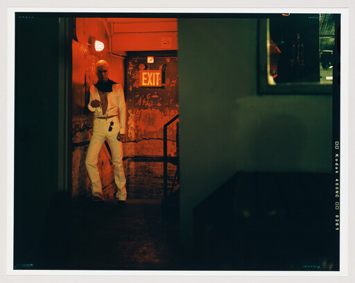 A person in white clothes stands under a red exit sign in a dark hallway.