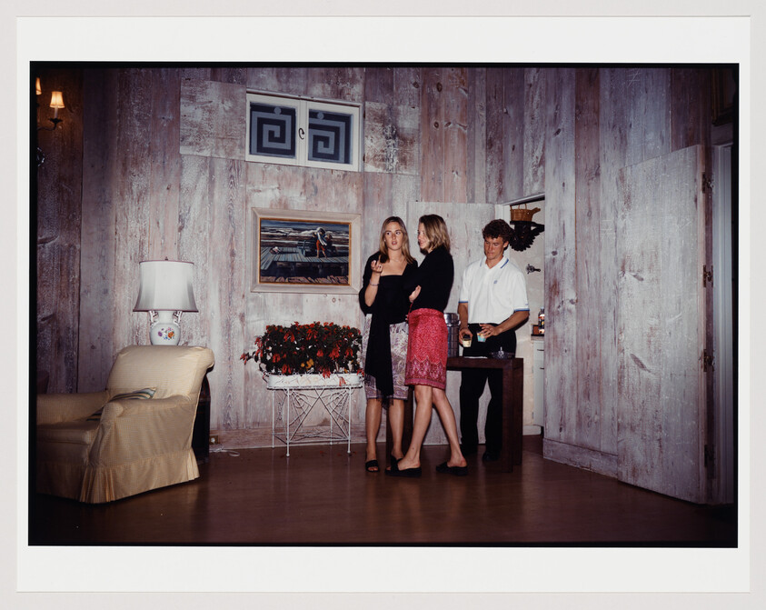 Two women chat while a man pours drinks at a small table in a wood-paneled room.