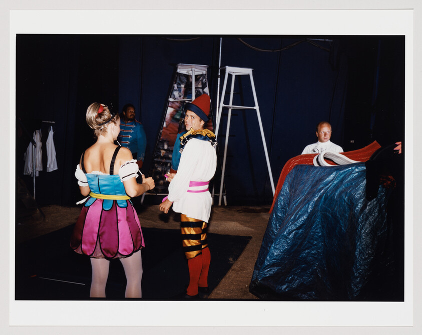 A photograph capturing a behind-the-scenes moment where two individuals in colorful, theatrical costumes are standing in a backstage area. The person on the left, facing away from the camera, wears a dress with a blue bodice and a pink and purple skirt, while the person on the right, facing the camera, is dressed in a white top with a blue and yellow collar, striped yellow and brown leggings, and red boots. In the background, another individual is partially obscured by a blue tarp, and a ladder is visible to the side.