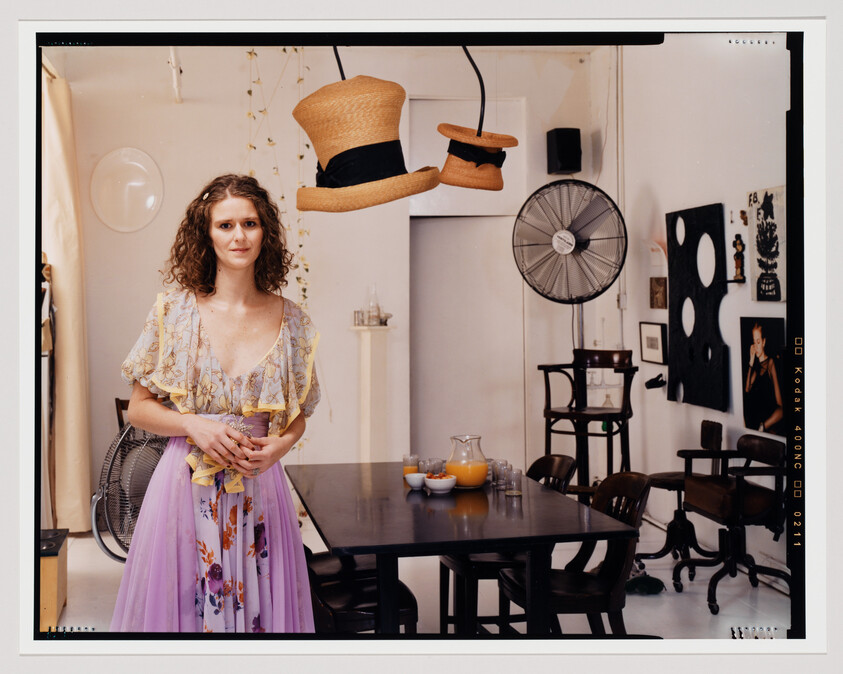 A woman with curly hair stands in the center of a room with eclectic decor, wearing a pastel floral dress. Above her, three hats are suspended in mid-air, and to the right, a vintage fan, a dark wooden table with chairs, and various wall decorations create an artistic and quirky atmosphere. The scene is framed within the borders of a film photograph, with film edge markings visible around the perimeter.