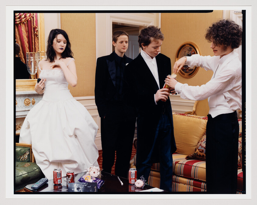 A candid scene in an elegant room with a young woman in a white dress looking at her phone, a young man in a black suit standing behind her, and another young man in a white shirt opening a bottle of champagne with a fourth person assisting. There are scattered items on the coffee table, including soda cans, a remote control, and a small bouquet.