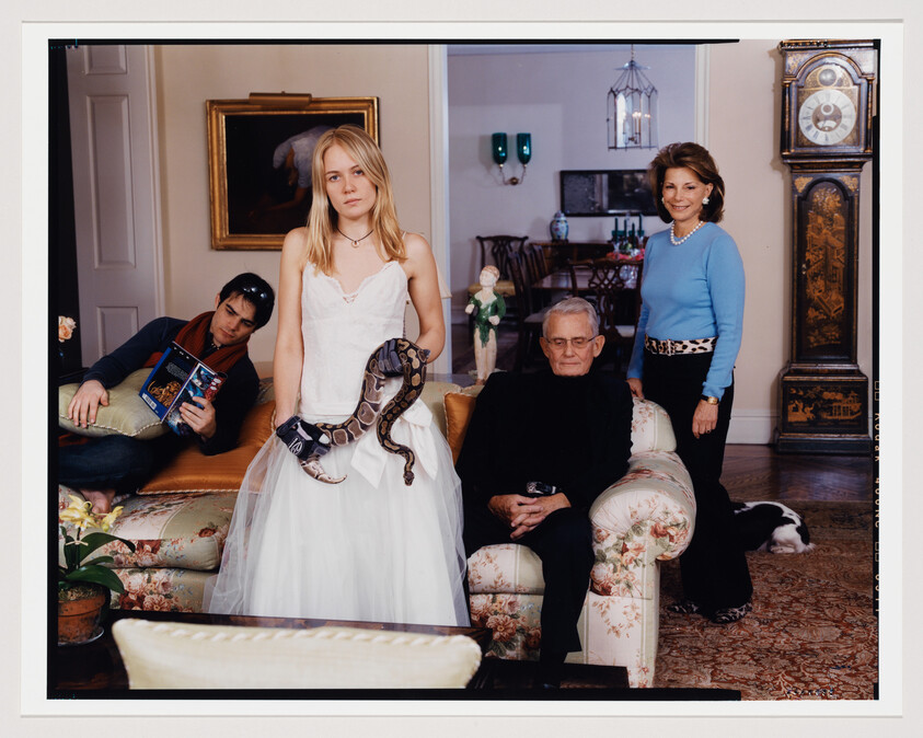 A staged family portrait in an elegant living room setting. A young woman in a white dress stands in the center holding a python around her shoulders. To her left, a young man lounges on a sofa, reading a book about magic, with a bored expression. To her right, an older man sits in an armchair, dressed in black and looking stern, while an older woman stands beside him, smiling at the camera, wearing a blue sweater and leopard print belt. A black and white dog lies on the floor, and the room is decorated with traditional furniture, paintings, and a tall antique clock.