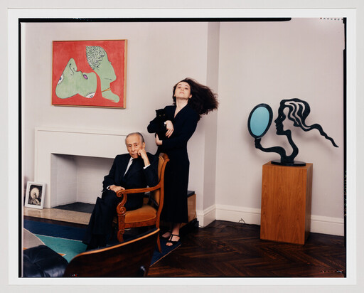 An elderly man sits in a chair while a woman behind him flips her hair.