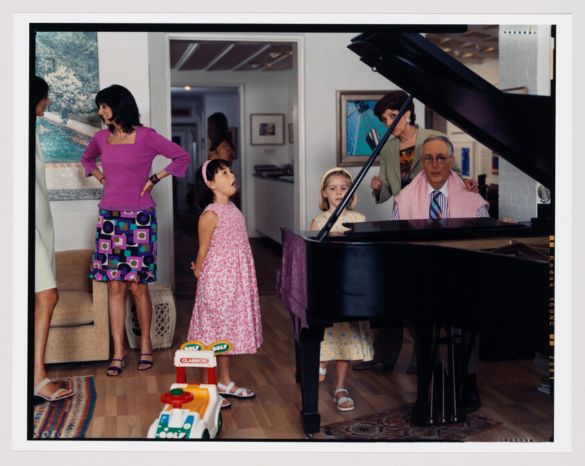 A family gathered around a grand piano in a cozy living room setting. A man is seated at the piano with sheet music in front of him, wearing a striped tie and a towel around his shoulders, while a woman stands behind him, appearing to adjust his hair or head. To the left, a woman in a purple top and patterned skirt converses with another woman partially visible in the background. In the foreground, a young girl in a pink dress looks up, seemingly in mid-conversation or song, and another young girl in a yellow dress stands by the piano, looking at the camera. Children's toys are scattered on the floor, adding a playful touch to the domestic scene.