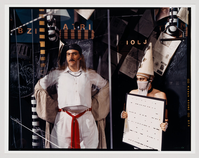 A staged photograph featuring two people in a surreal setting. The person on the left is wearing a white shirt, beige pants, and a red sash, with a clear plastic sheet in front of their face, behind which various scribbles are visible. The person on the right is wearing a white shirt and a paper cone hat, with a mask covering their mouth, and is holding a large clipboard with musical notes. The background is chaotic with newspaper clippings and abstract shapes.