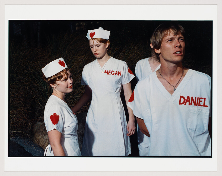 Three individuals in white nurse uniforms with red hearts and name tags stand against a backdrop of tall grass. The two women wear traditional nurse's caps with a red heart, while the man in the center looks away from the camera. The name tags read "MEGAN" and "DANIEL" respectively.