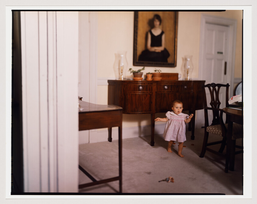 A toddler in a pink dress takes unsteady steps across a dining room rug near furniture.