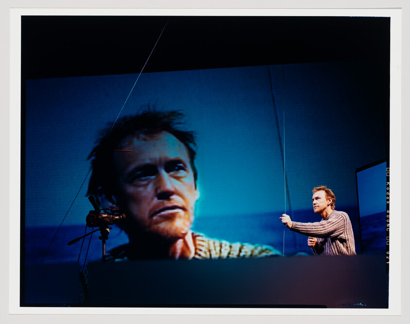 A man pulls a rope while a large projected portrait of his face fills the screen.