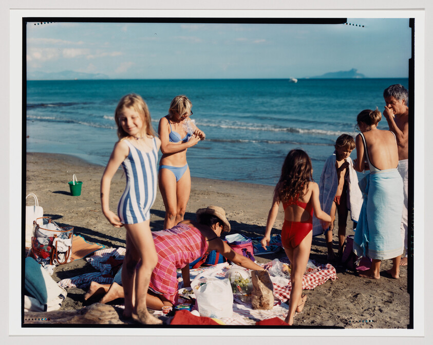 Children and adults gather on a sandy beach unpacking picnic items and preparing to swim.