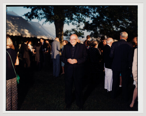Smiling priest in black clerical suit stands with hands clasped at an outdoor evening gathering.