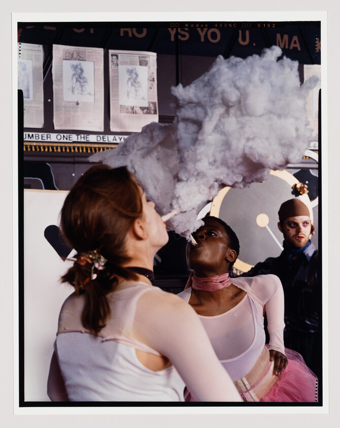A person exhales a large cloud of smoke towards another person's face, with a bystander looking on, against a backdrop of posters.