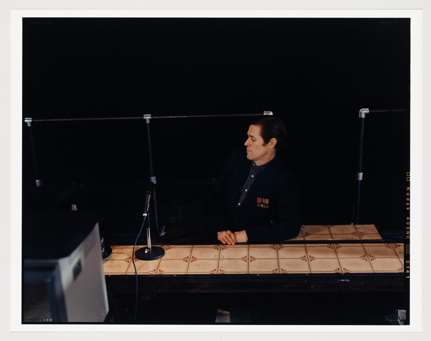 A man in a dark suit with medals on his chest sits at a table with a patterned surface, looking to the side. In front of him is a microphone stand, and the background is predominantly dark, with the top edge of a monitor visible in the lower left corner. The image has a white border with markings typical of a film contact sheet.