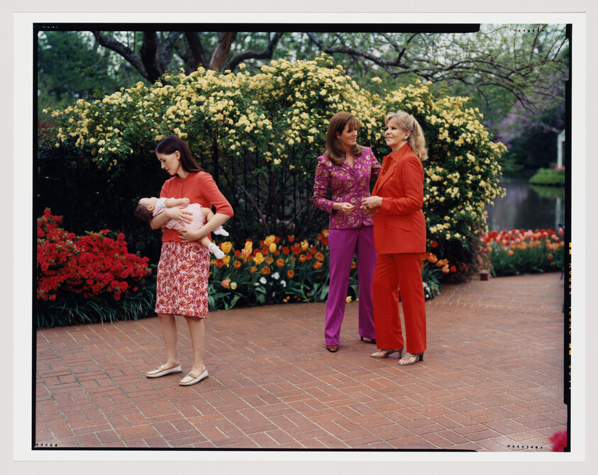 Three women stand in a vibrant garden with blooming flowers. On the left, a woman in a red top and patterned skirt cradles a sleeping baby. In the center, two women engage in conversation, one dressed in a purple patterned suit and the other in a red suit. The background features lush yellow rose bushes and a variety of tulips, with a calm pond visible in the distance.