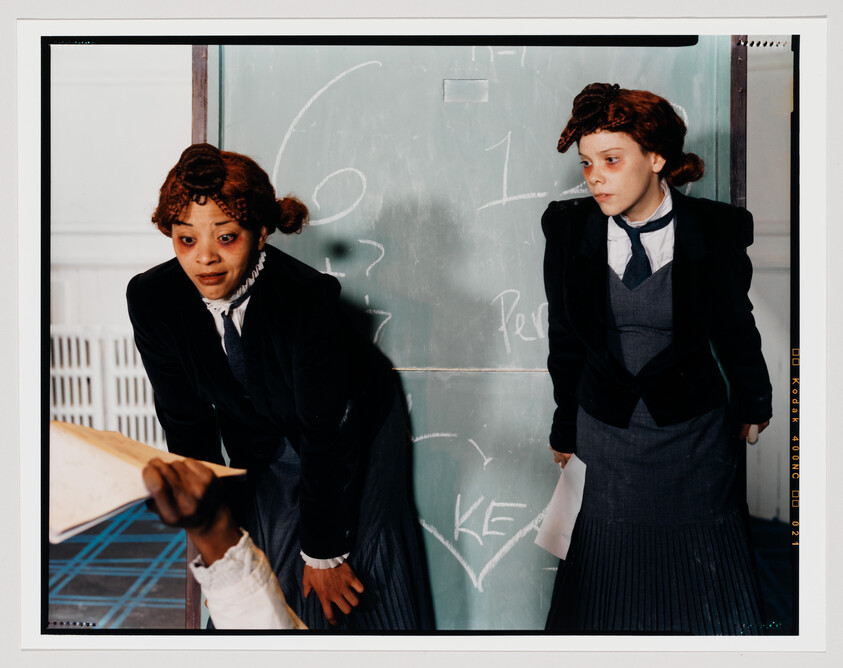 Two girls in old-fashioned school uniforms stand by a chalkboard while one reaches for a paper.