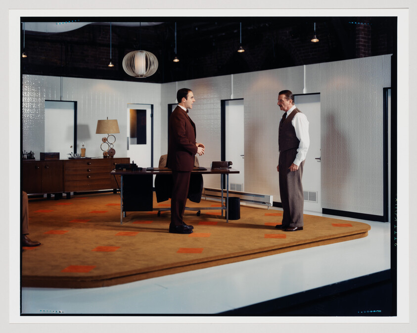 Two men in a vintage office stand facing each other near a desk, appearing to talk.
