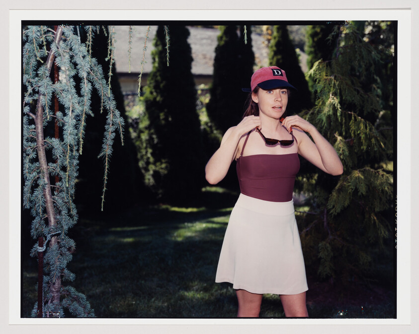 Young woman in a red cap adjusts sunglasses around her neck while standing in a shady garden.