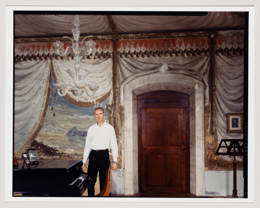 A man in a white shirt stands by a piano in an ornate room with a chandelier.