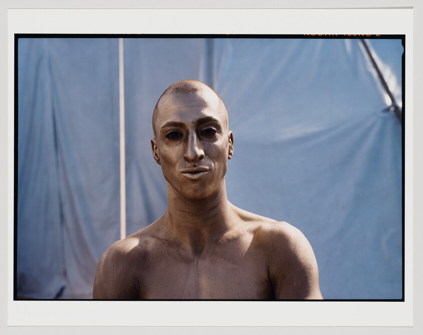 A photograph of a shirtless person with a metallic gold painted face and upper body, posing in front of a plain backdrop with a slight crease visible. The person has a neutral expression and is looking directly at the camera. The photo includes a white border with film information visible at the top edge.