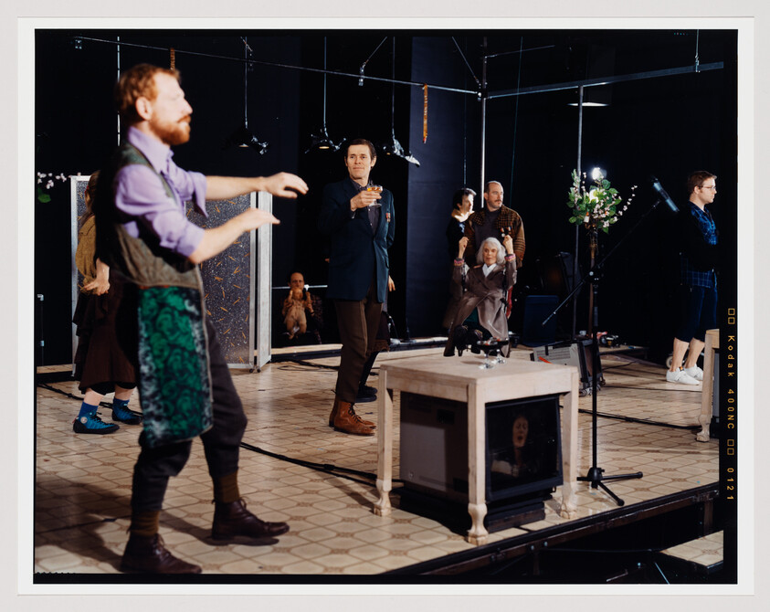 A dynamic scene on a theater stage with multiple actors and props. In the foreground, a man with a beard is gesturing with his arm extended, wearing a purple shirt and a green patterned apron. In the middle, a man in a suit holds a drink, looking off to the side. Behind him, various performers are scattered, including one seated in a wheelchair. The stage is adorned with mirrors, lights, and a glass table with a vase of flowers. The setting suggests a rehearsal or a performance in progress.