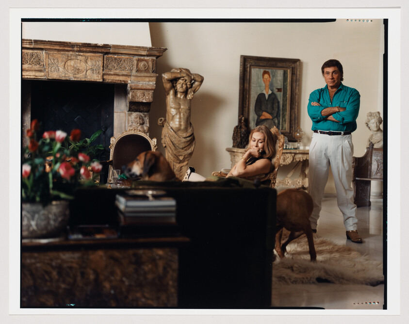 A man stands with arms crossed while a woman sits nearby in an ornate living room.